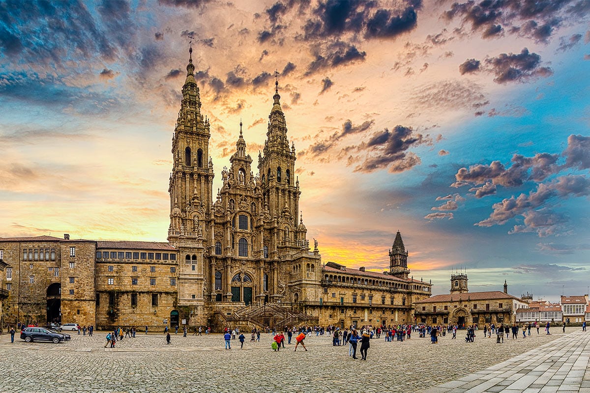A large cathedral with two tall towers stands in a cobblestone square, where groups of people gather beneath a dramatic, colorful sunset—capturing the spirit of the Camino de Santiago during Holy Year.