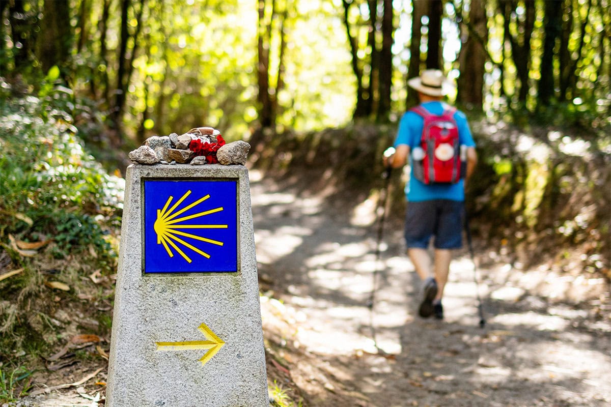 A Camino de Santiago trail marker with a yellow arrow and shell symbol in the foreground; a hiker with sticks and a red backpack walks on a wooded path, experiencing the Camino do Santiago during Holy Year.