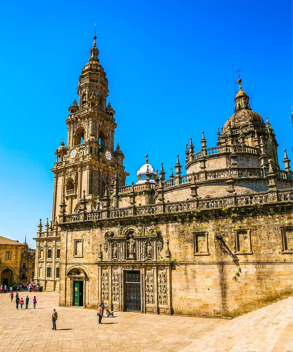 The exterior of a historic stone cathedral, a renowned stop on the Camino de Santiago during Holy Year, features ornate towers and detailed carvings beneath a clear blue sky, as people stroll through the lively plaza in front.
