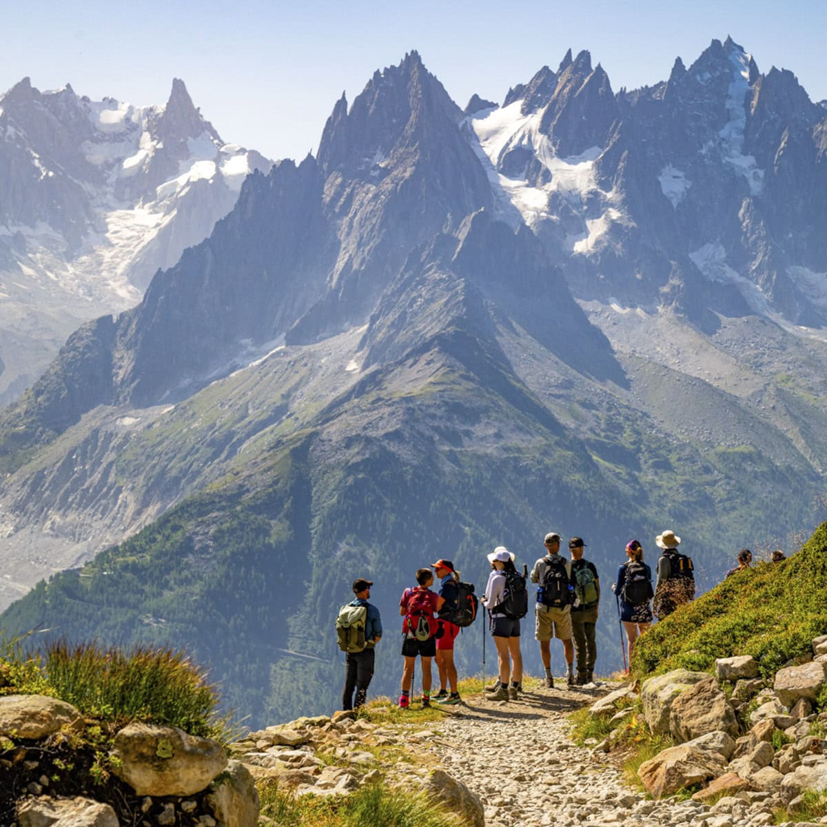 A group of hikers stands on a rocky mountain trail along the famous hiking Tour du Mont Blanc, with jagged, snow-capped peaks and green slopes in the background under a clear sky.