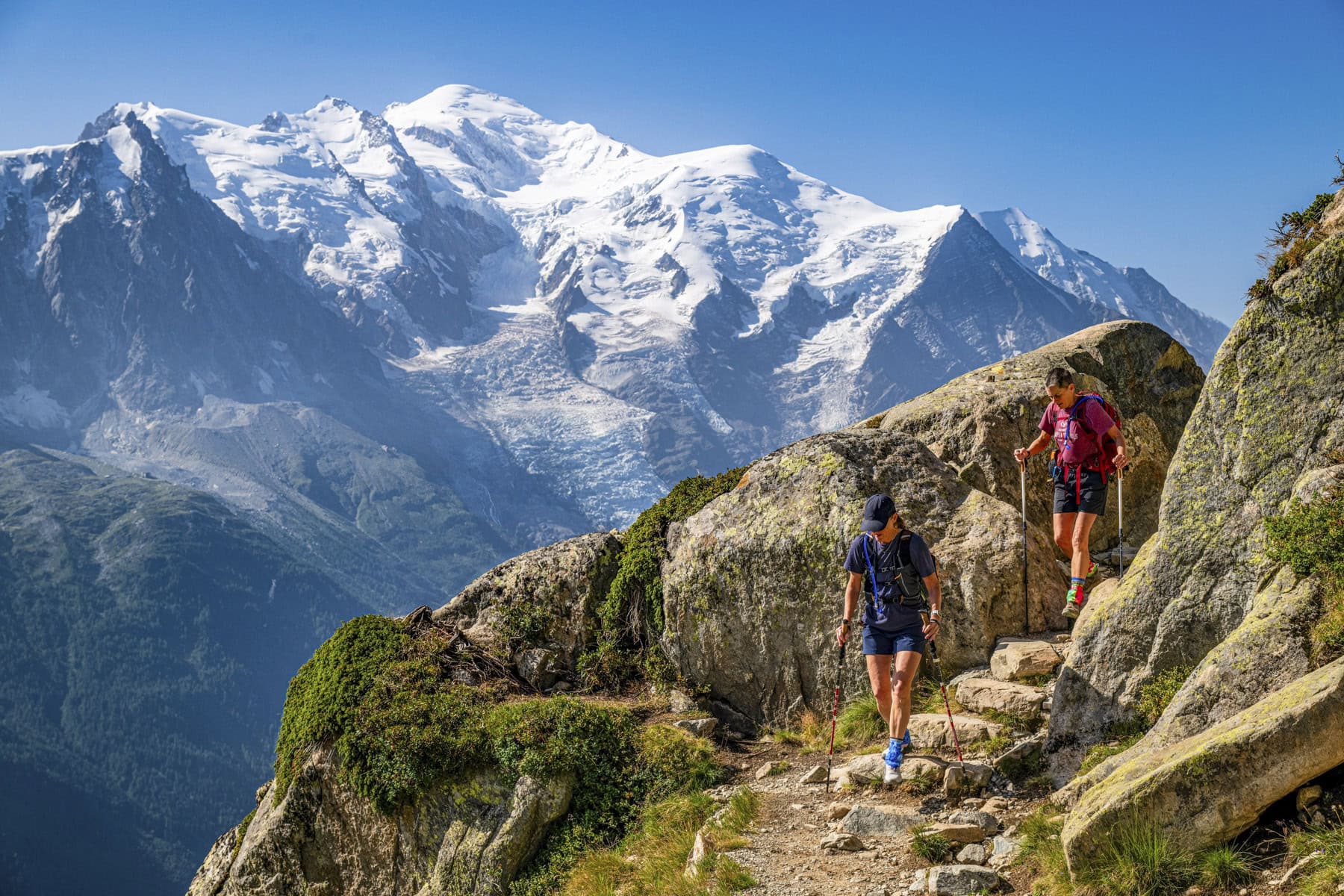 Two hikers walk up a rocky mountain trail with snow-capped peaks and a clear blue sky in the background, exploring the breathtaking scenery of the Tour du Mont Blanc.