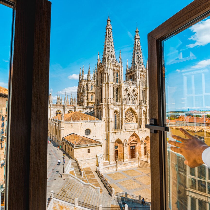 View through an open window of a large Gothic cathedral with tall spires and intricate architecture under a clear blue sky, evoking the spirit of Camino de Santiago during Holy Year. A hand is visible opening the window.