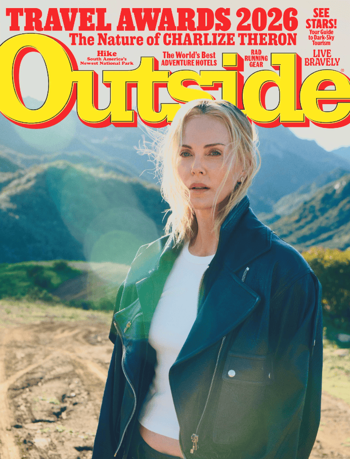 A woman in a black leather jacket stands on a dirt path with green mountains behind her, featured on the 2026 cover of Outside Magazine’s Best Adventure Travel Companies issue.