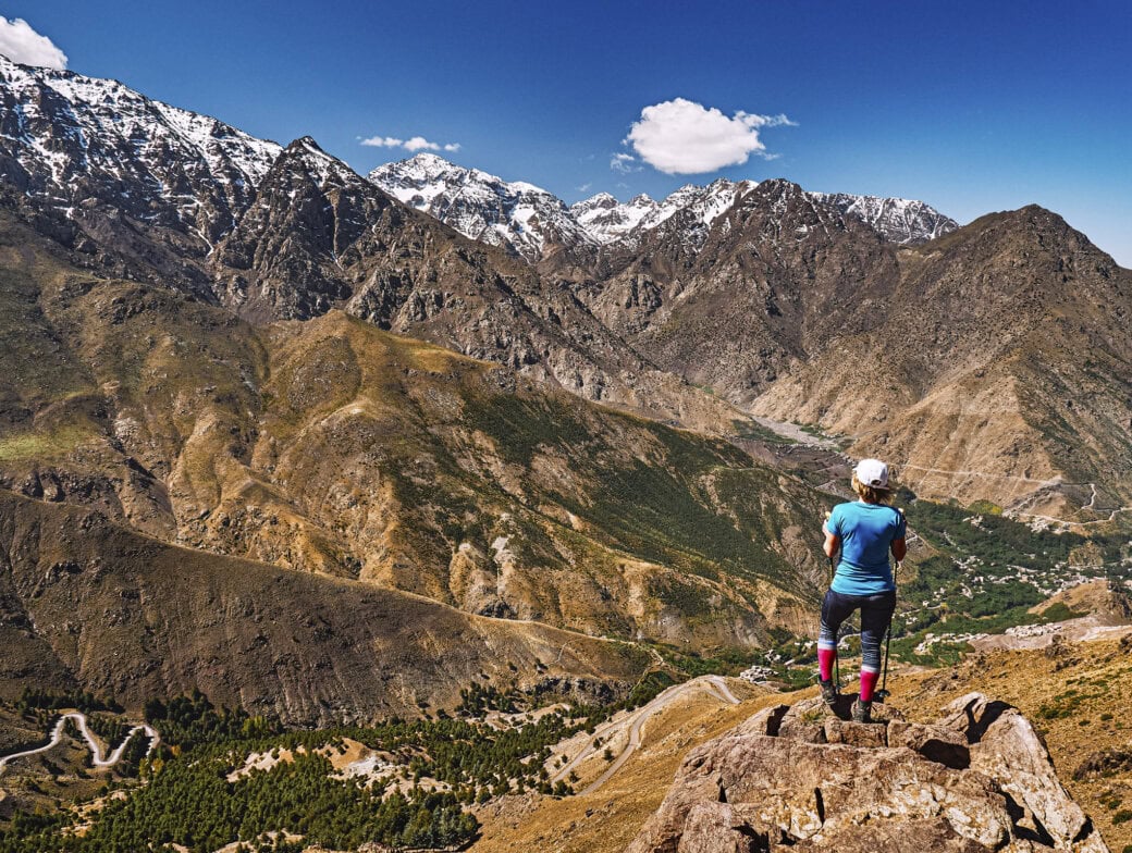 Person standing on a rocky outcrop in Morocco overlooks a vast mountain landscape with snow-capped peaks and winding roads under a clear blue sky, capturing the breathtaking views during the best time to visit.