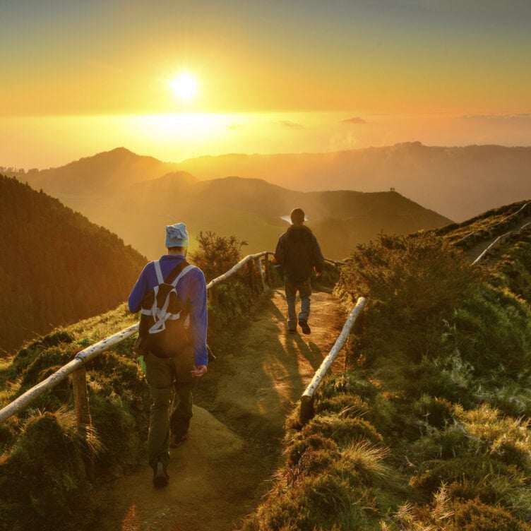 Two people hike a dirt path along a mountain ridge at sunrise—one of the unforgettable Azores things to do—while sunlight casts long shadows and illuminates the breathtaking landscape.
