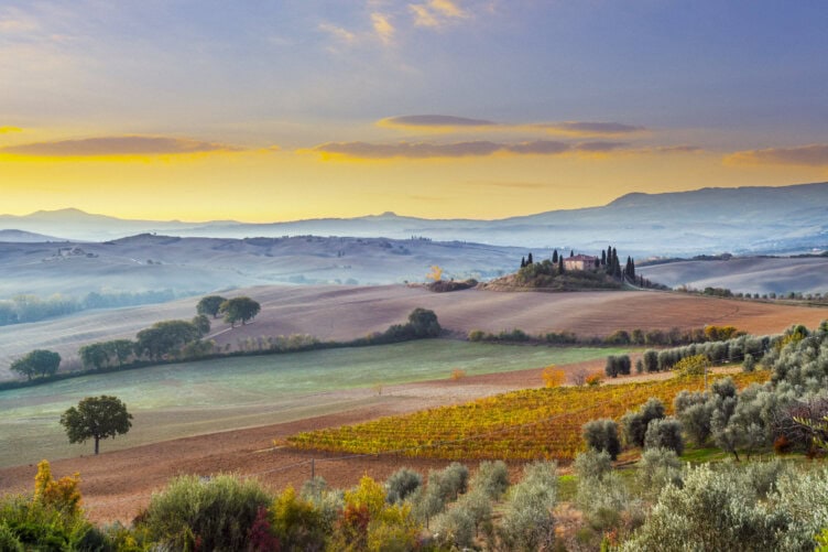 Rolling hills with patchwork fields, trees, and a farmhouse atop a hill under a colorful sky at sunrise or sunset—the best time to visit Italy for scenes like this.