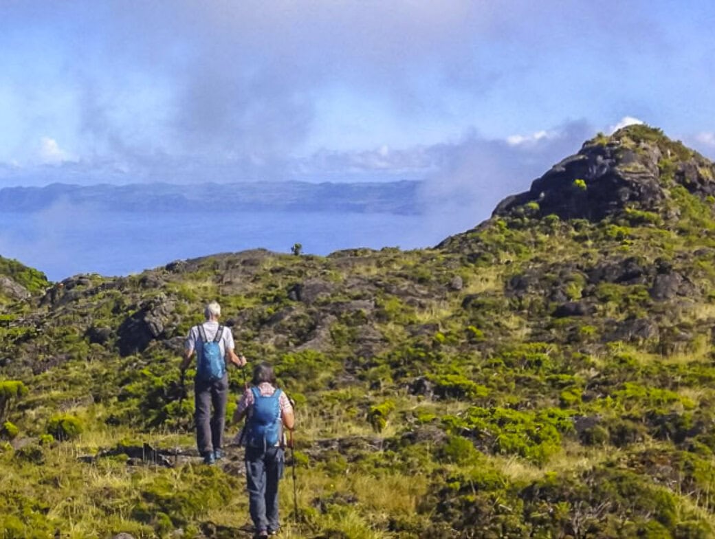 Two hikers with backpacks walk along a grassy, rocky trail in the Azores—one of the top Azores things to do—with a hill to the right and distant views of water and mountains under a partly cloudy sky.