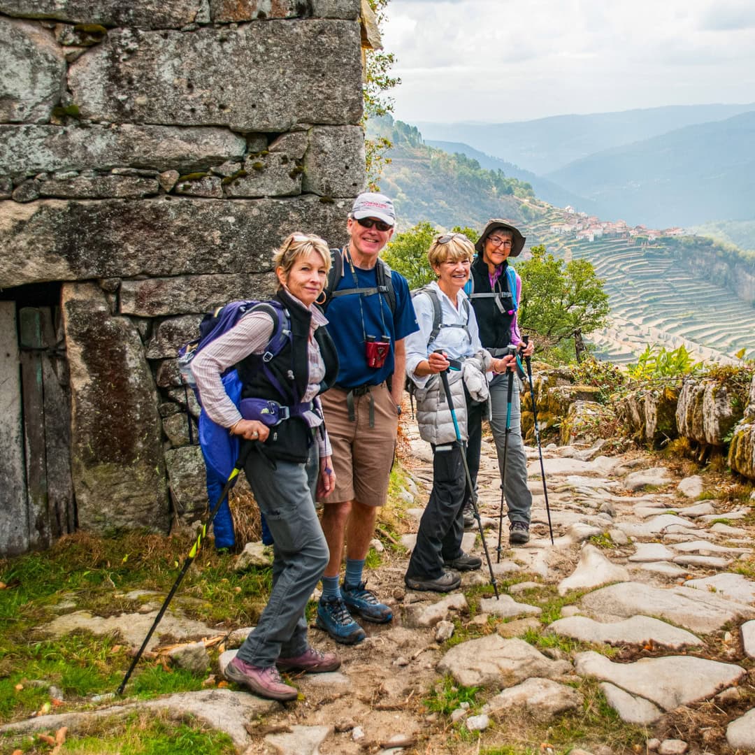Four hikers with trekking poles stand on a rocky trail beside an old stone building, taking in the terraced hills and distant village—an unforgettable Portugal hiking experience.