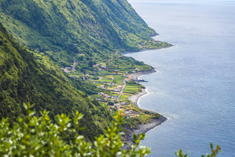 A coastal village with houses and green fields lies between steep, forested cliffs and the blue ocean under a clear sky—one of the many picturesque scenes to discover among Azores things to do.