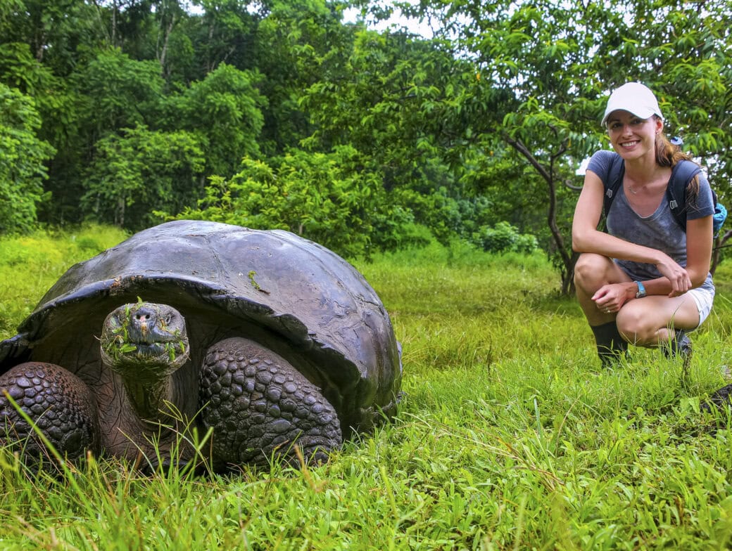A large tortoise sits on grass near a smiling woman wearing a white cap, with dense green foliage in the background—capturing the magic of wildlife during the Best Time to Visit the Galapagos.