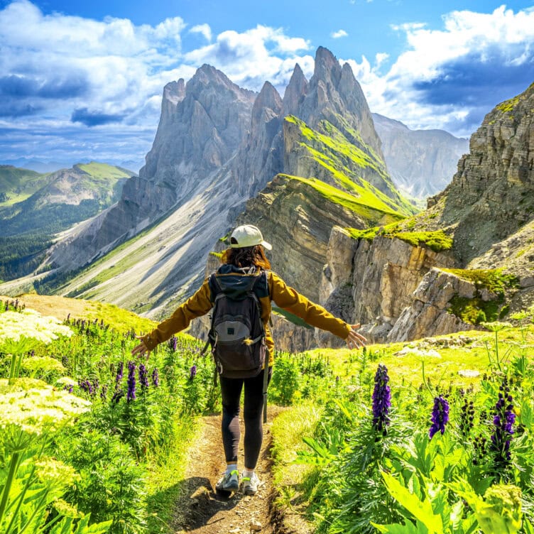 A person with a backpack walks along a mountain trail surrounded by wildflowers, with jagged peaks and green hills in the background under a partly cloudy sky—the scene evokes the best time to visit Italy’s breathtaking landscapes.