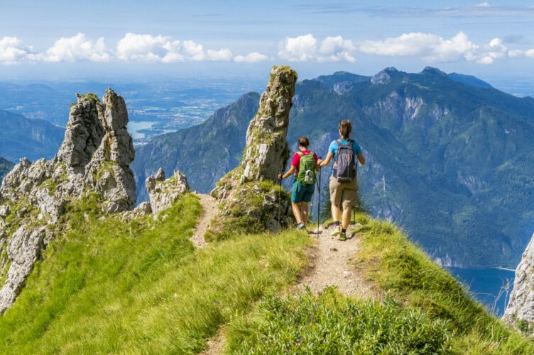 Two hikers with backpacks walk along a grassy mountain ridge, surrounded by rocky peaks and overlooking a valley and distant lake under a partly cloudy sky—the perfect scene to experience during the best time to visit Italy.