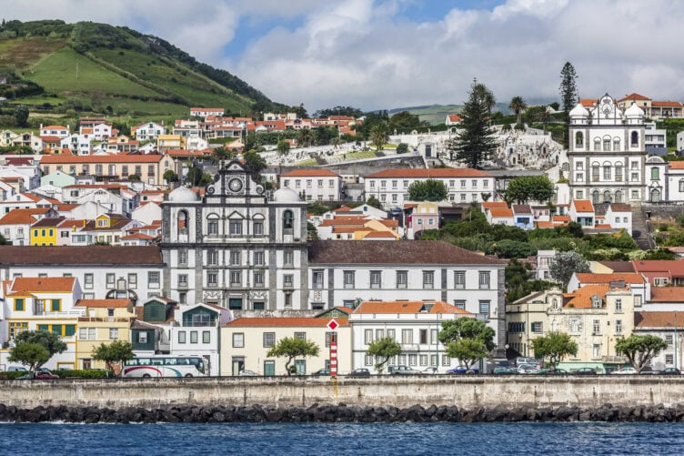Colorful buildings with red-tiled roofs line a coastal town in the Azores, with hills behind and a baroque-style church prominently visible—a classic scene and one of the top Azores things to do for travelers.
