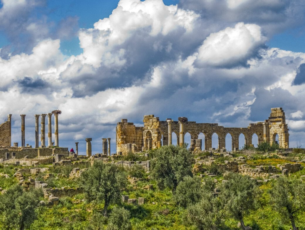 Ancient stone ruins with columns and arches stand on a grassy landscape under a cloudy blue sky, evoking the timeless beauty of Morocco. Discover the site’s charm and history—explore to find the best time to visit for an unforgettable experience.