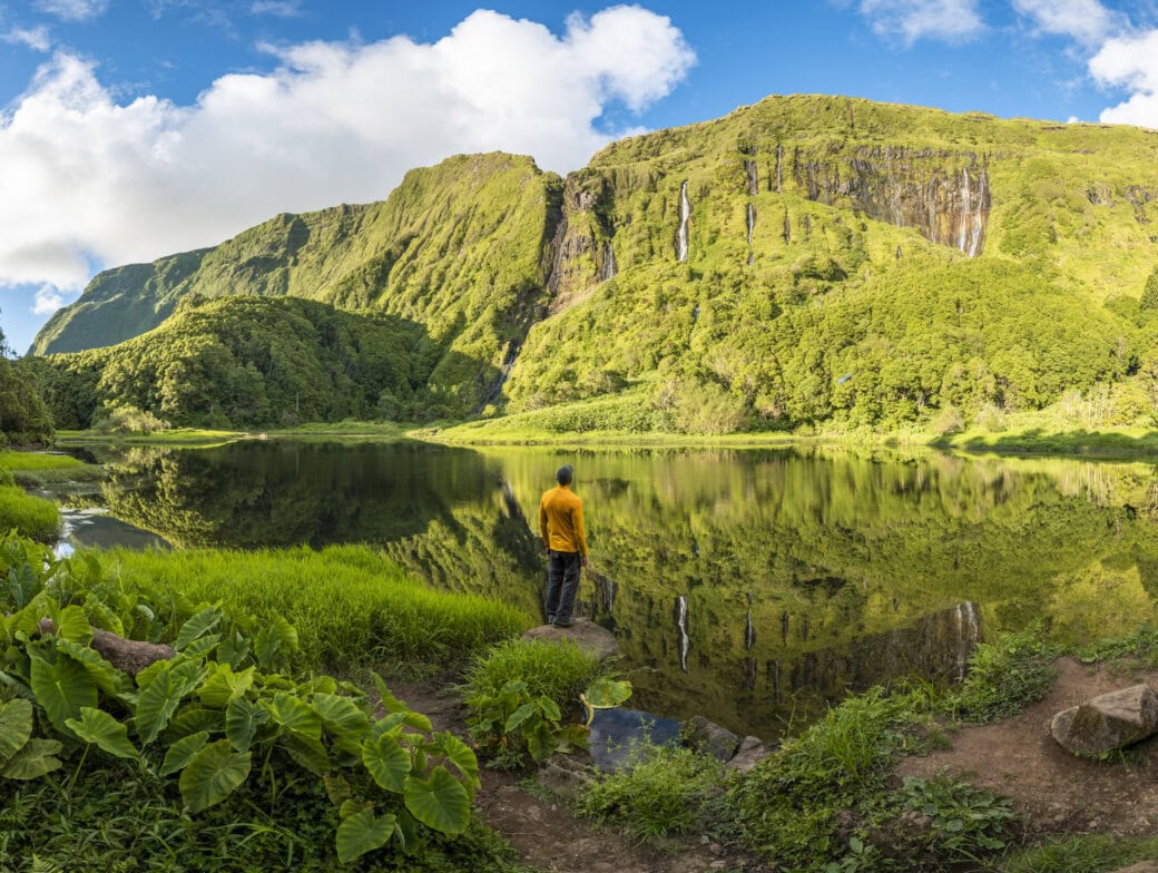 A person in a yellow jacket stands by a calm lake surrounded by green mountains and lush vegetation under a blue sky with scattered clouds, capturing the beauty of Azores things to do.
