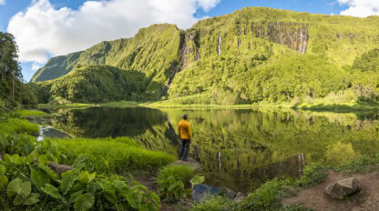 A person in a yellow jacket stands by a calm lake surrounded by green mountains and lush vegetation under a blue sky with scattered clouds, capturing the beauty of Azores things to do.