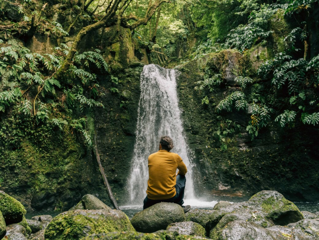 A person in a yellow sweater sits on rocks facing a small waterfall, surrounded by lush green trees and foliage—a serene moment among the many Azores things to do.