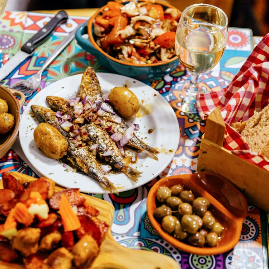 A table set with grilled sardines, potatoes, sliced bread, olives, a tomato salad, cured meats, and a glass of white wine—taste the real Portugal after a day of hiking Portugal with an expert guide.