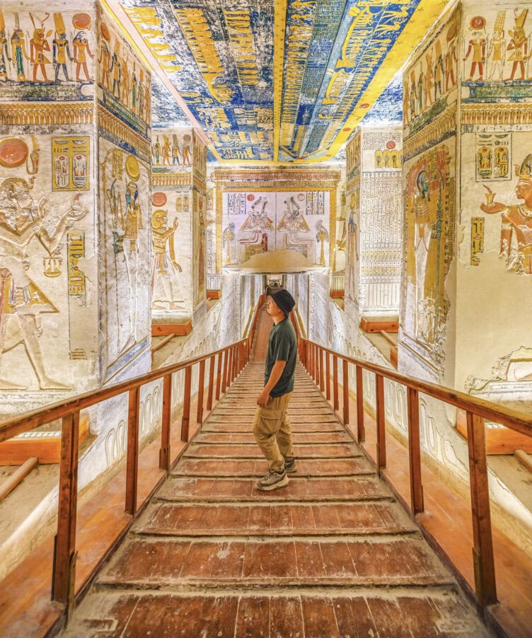 A person stands on a wooden walkway inside an ancient Egyptian tomb, surrounded by colorful hieroglyphics and detailed wall paintings—a captivating glimpse into what egypt tours have to offer.