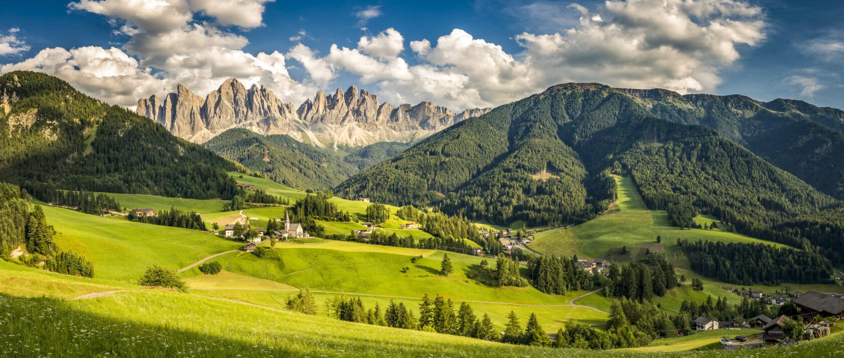 A panoramic view of a lush green valley with scattered houses, surrounded by forested hills and the jagged peaks described in many Dolomites itineraries, all beneath a partly cloudy sky.