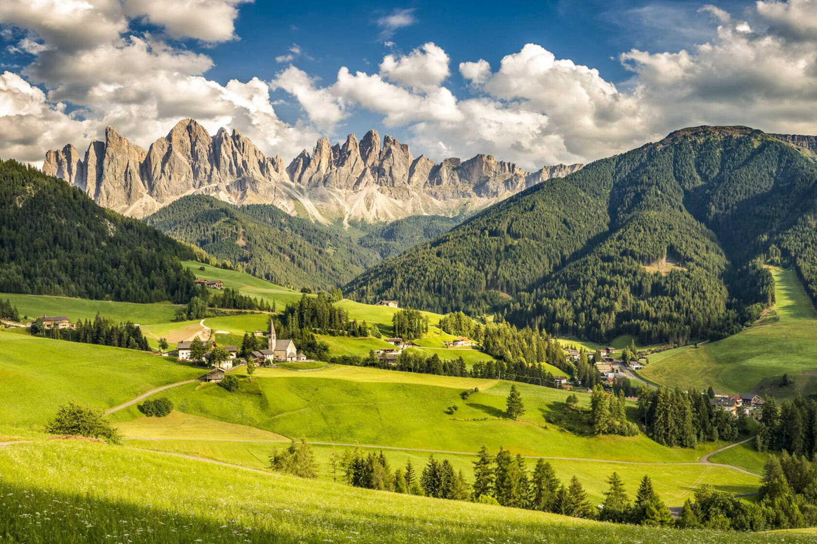 A small village with a church sits in a green valley surrounded by rolling hills and forest, with rugged mountains in the background under a partly cloudy sky.