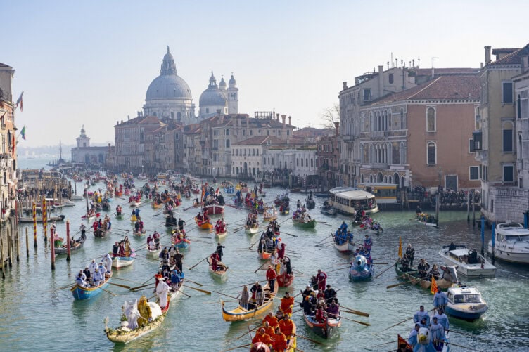 A large number of colorful rowboats and gondolas fill a Venice canal, with crowds of people aboard and historic buildings and domes visible in the background—capturing the vibrant scene during the best time to visit Italy.