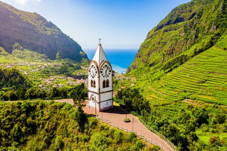 A white church tower with a clock stands among green hills, overlooking a valley and the ocean under a clear blue sky—an iconic scene featured in any Madeira travel guide.