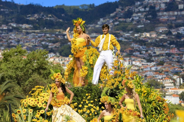 Four people in yellow outfits and floral decorations stand on a flower-covered parade float, set against a scenic hilly town and blue sky—an enchanting scene straight out of a Madeira travel guide.