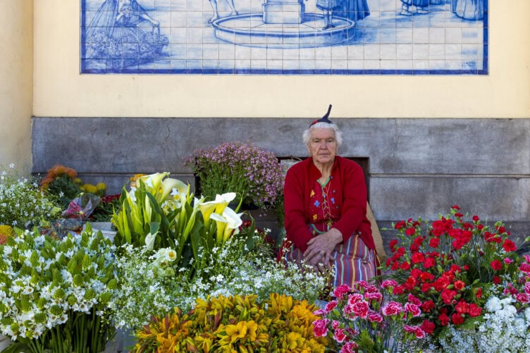 An elderly woman wearing a red sweater sits among colorful flowers at an outdoor market, with a decorative blue tile mural behind her—a charming scene straight from any Madeira travel guide.