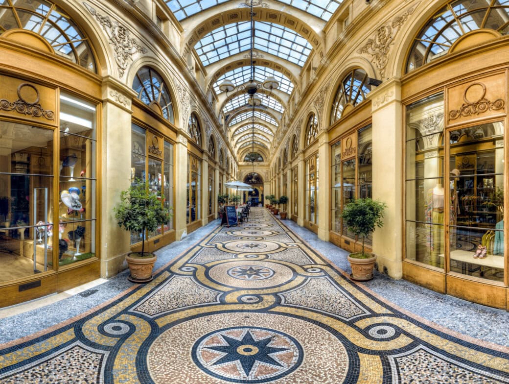 Ornate indoor shopping arcade with arched glass ceiling, mosaic tile floor, potted plants, and boutique storefronts on both sides—a must-see Paris attraction for any Paris travel itinerary.