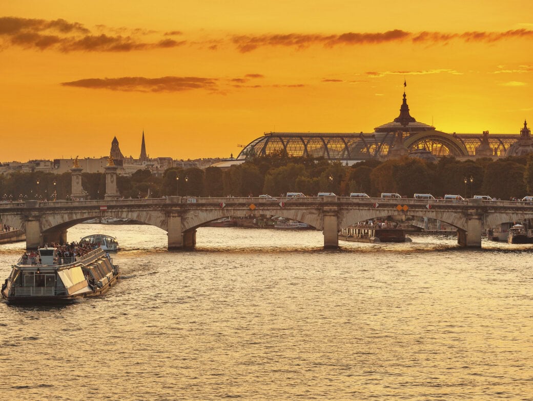 A riverboat glides on the Seine beneath a stone bridge at sunset, with historic buildings and the Grand Palais in view—an unforgettable experience among top Paris attractions.