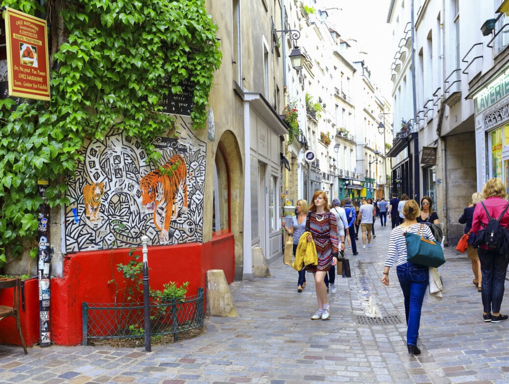 People walk along a cobblestone street lined with shops, graffiti art, greenery, and outdoor seating in a lively urban neighborhood—one of the must-see Paris attractions for anyone interested in Paris travel.