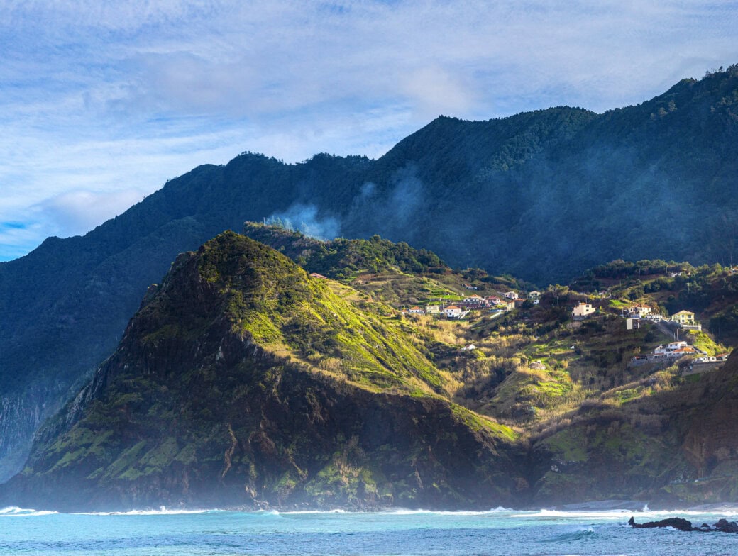 A coastal mountain landscape featured in the Madeira travel guide, with scattered houses on green hillsides, mist rising, and waves crashing at the shore under a partly cloudy sky.