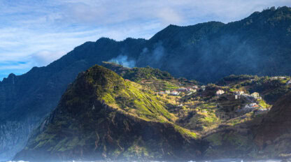 A coastal mountain landscape featured in the Madeira travel guide, with scattered houses on green hillsides, mist rising, and waves crashing at the shore under a partly cloudy sky.