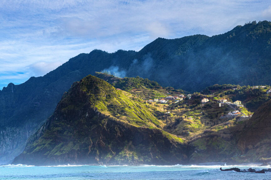 A coastal mountain landscape featured in the Madeira travel guide, with scattered houses on green hillsides, mist rising, and waves crashing at the shore under a partly cloudy sky.