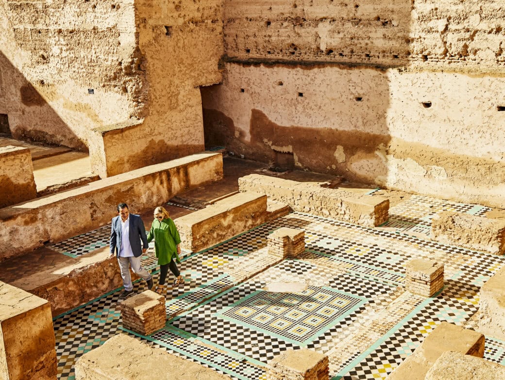 Two people walk across a patterned mosaic floor among ancient stone ruins in Morocco, with high textured walls and sunlight casting shadows—the best time to visit for exploring timeless architecture.