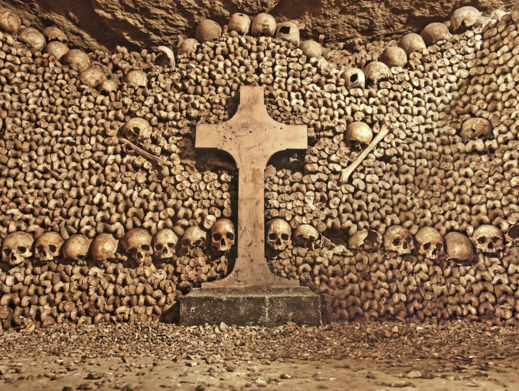 A stone cross stands before walls of neatly stacked human skulls and bones in an underground crypt—one of the most unusual Paris attractions for those seeking unique Things to Do in Paris.