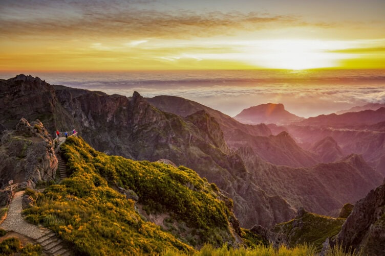A mountain landscape at sunrise with a pathway and people standing on a viewpoint, overlooking green peaks and a sea of clouds below—a perfect scene for any Madeira travel guide.