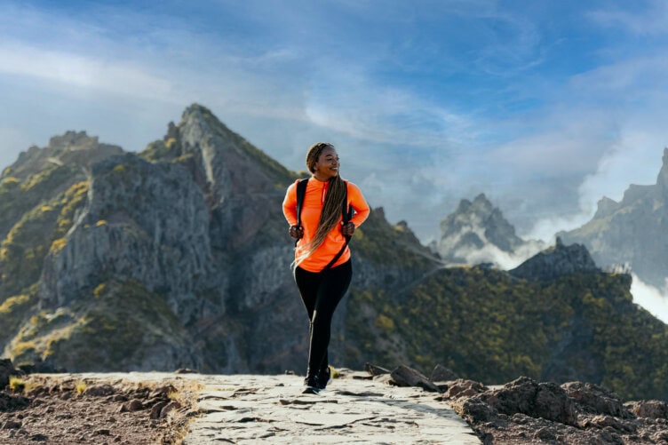 A person wearing an orange jacket and backpack walks on a stone path in a mountainous area under a blue sky with clouds, capturing the spirit of adventure found in any Madeira travel guide.