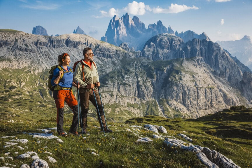 Two hikers with backpacks and trekking poles stand on a grassy mountain ridge in the Dolomites, overlooking jagged peaks under a blue sky—a classic scene found in any Dolomites travel guide when exploring the best time to visit.