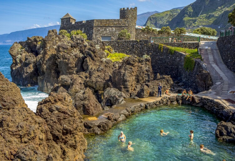 People swim in a natural pool surrounded by volcanic rock, with a stone building and mountains in the background under a clear blue sky—a must-see spot featured in any Madeira travel guide.