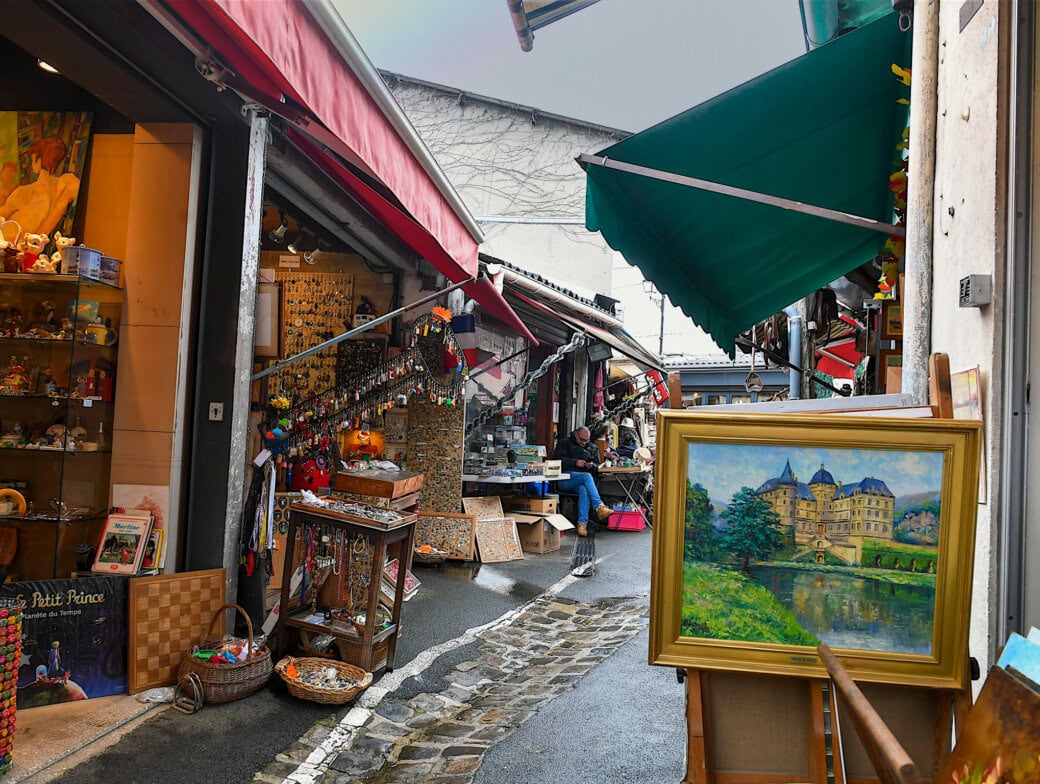 Narrow outdoor market street with art, paintings, and souvenirs displayed in shops on both sides under colorful awnings—one of the charming Paris attractions to explore during your Paris travel.