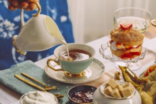 A hand pours milk into a teacup on a table set with gold-trimmed china, cream-filled pastry with strawberries, sugar cubes, and preserves—an elegant scene reminiscent of classic London England afternoon teas.