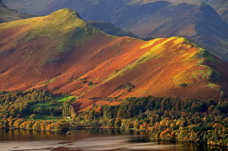 Rolling hills with green and orange foliage, trees at the base, and a lake in the foreground under soft sunlight evoke the tranquil charm of England countryside regions.