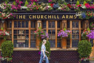 A person walks past The Churchill Arms pub in London, which has wooden windows and is decorated with numerous colorful flower arrangements—a classic spot among Things to Do in London.