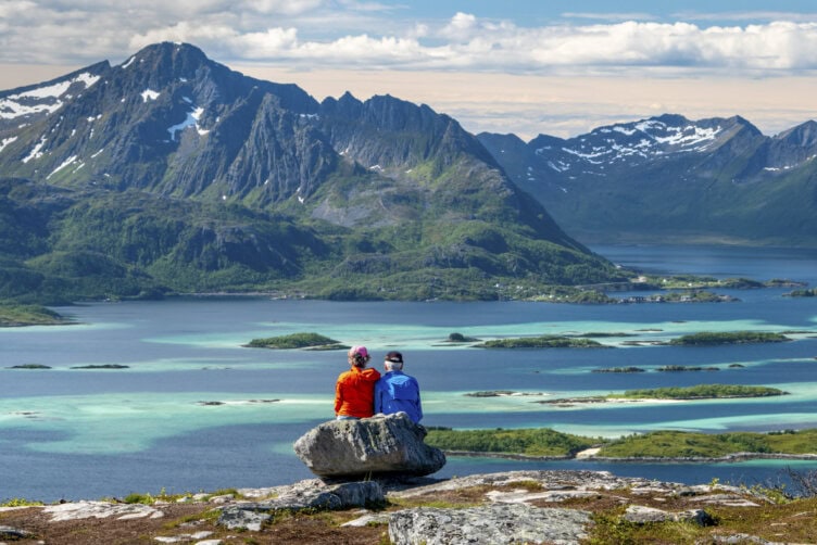 Two people sit on a rock overlooking turquoise water, small islands, and snow-capped mountains under a partly cloudy sky.