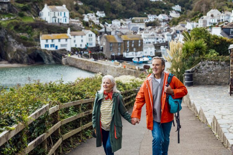An older couple walks hand in hand along a coastal path in a seaside village, surrounded by houses, greenery, and the ocean, capturing the charm of England countryside regions.