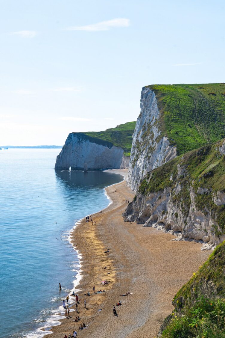 A sandy beach with people relaxing beside tall white cliffs and green hills, typical of England countryside regions, next to a calm blue sea under a clear sky.