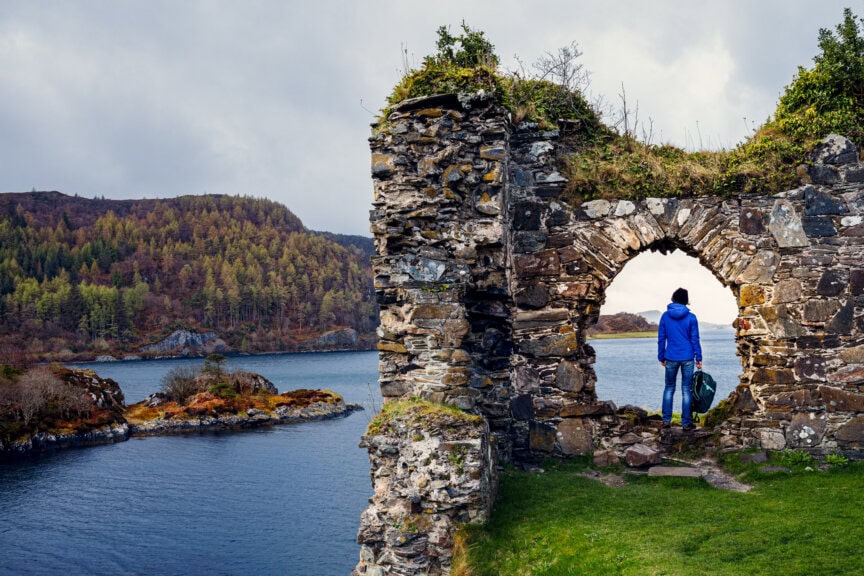 A person with a backpack stands in a stone archway overlooking a lake and wooded hills under a cloudy Scotland sky, capturing the best time to visit this breathtaking landscape.