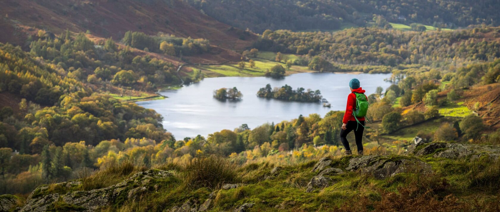A person in outdoor gear stands on a hillside overlooking a scenic lake surrounded by trees and rolling hills, capturing the beauty of England countryside regions in autumn.